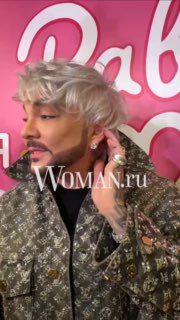 Крупный план Филиппа Киркорова на розовом баннере премьеры «Равиоли Оли», артист поправляет серьгу, на заднем плане фрагмент логотипа Woman.ru