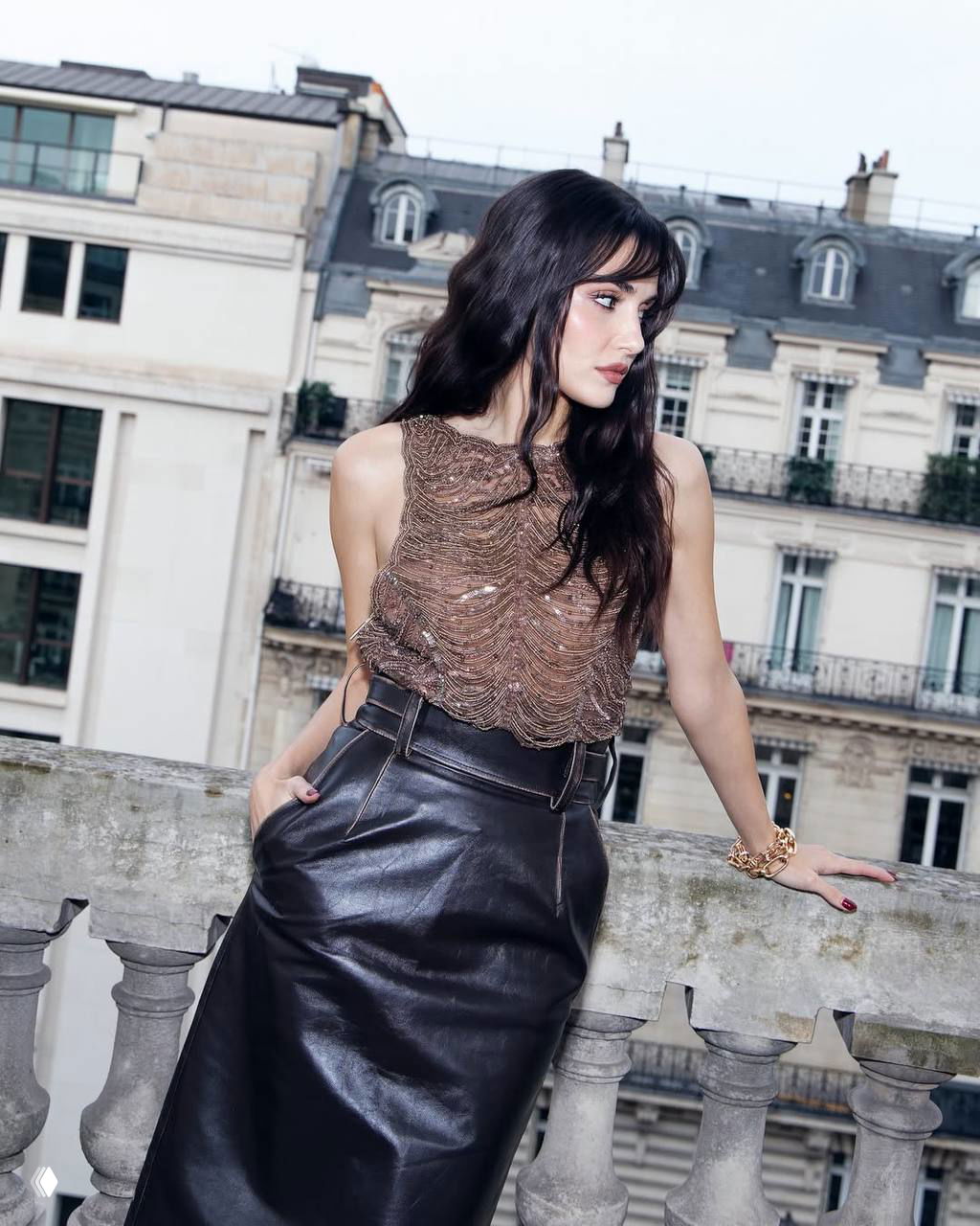 Актриса в кожаной юбке и прозрачном коричневом топе позирует на балконе с видом на исторические здания — фото с московского показа.