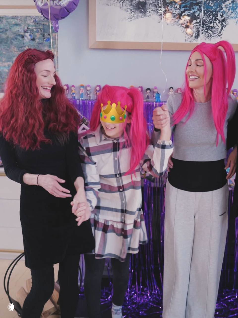 Три поколения женщин держатся за руки и улыбаются перед фиолетовой праздничной растяжкой, центральное лицо ребёнка закрыто стикером для приватности.