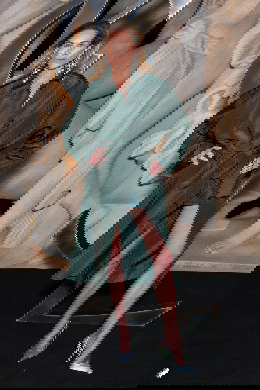 Модель в длинном зелёном плаще с завязками и высоких туфлях позирует у фирменной инсталляции Saint Laurent на показе в Париже.