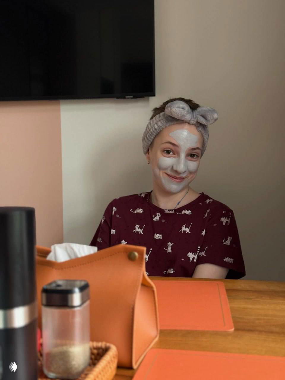 Подросток в повязке для волос с косметической маской на лице сидит за столом в домашней обстановке, на столе видны косметика и аксессуары.