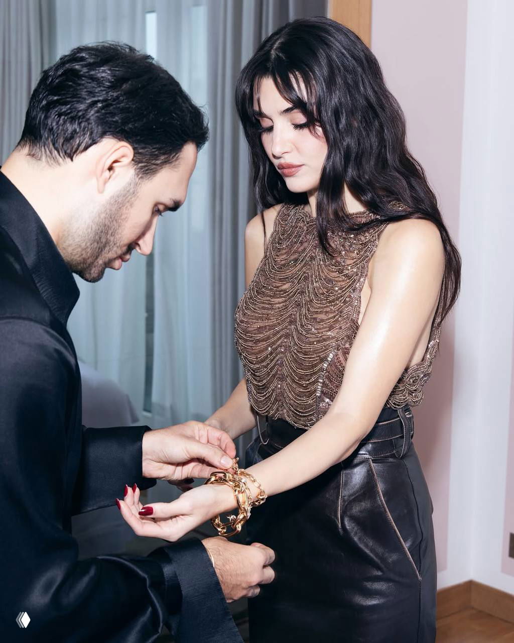 Кадр с места мероприятия: актриса получает украшение на руку от гостя, в наряде с цепочными браслетами и элегантным маникюром.
