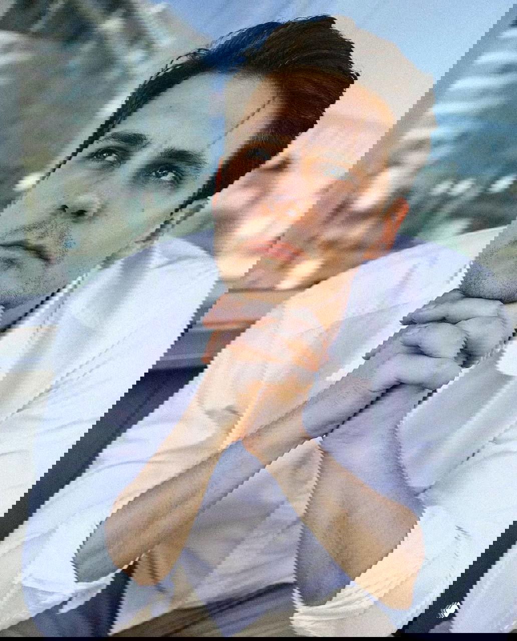 Крупный портрет Милоша Биковича в белой рубашке, задумчивый взгляд вверх, городская размытая архитектура на заднем плане.