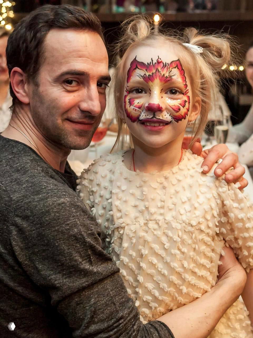 Мужчина обнимает ребёнка с раскрашенным аквагримом на лице: тёплая семейная сцена в помещении, праздничная атмосфера.