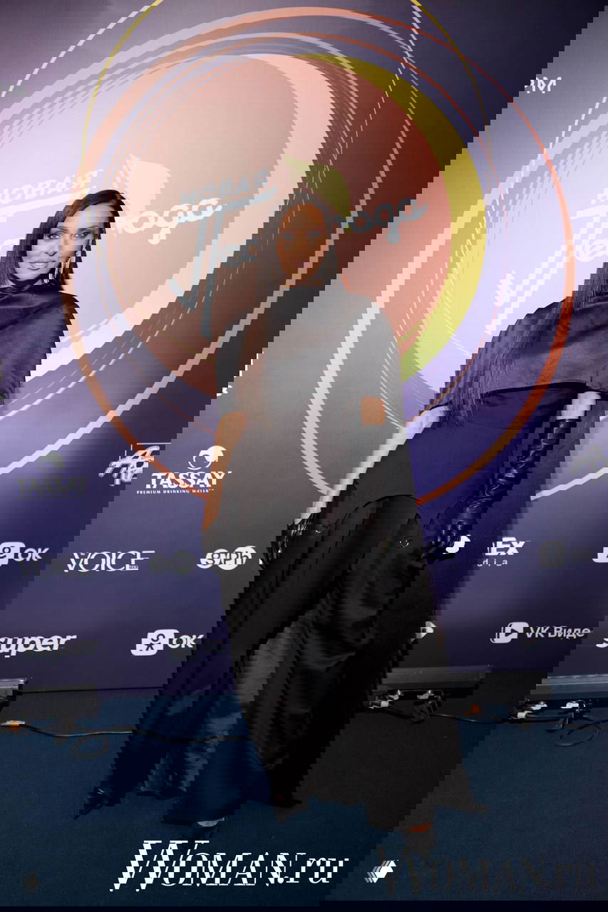 Артистка в чёрном длинном платье и перчатках позирует на фоне баннера фестиваля «Новая Песня года» — крупный портрет, полный рост.