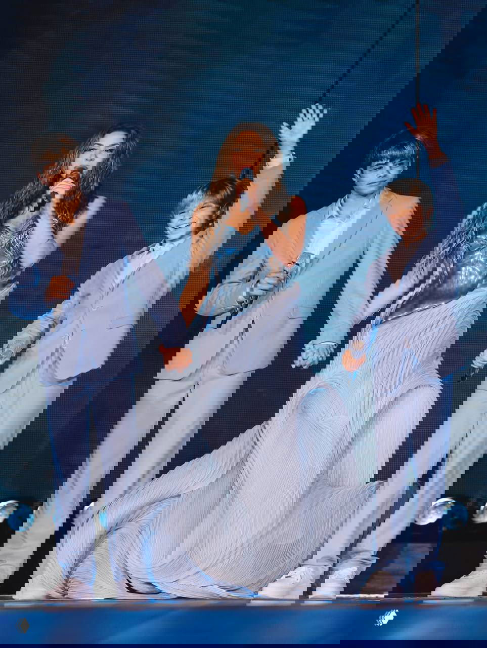 Шакира держит за руки сыновей на сцене; мальчики улыбаются и машут публике, все в голубой сценической гамме и элегантных костюмах.