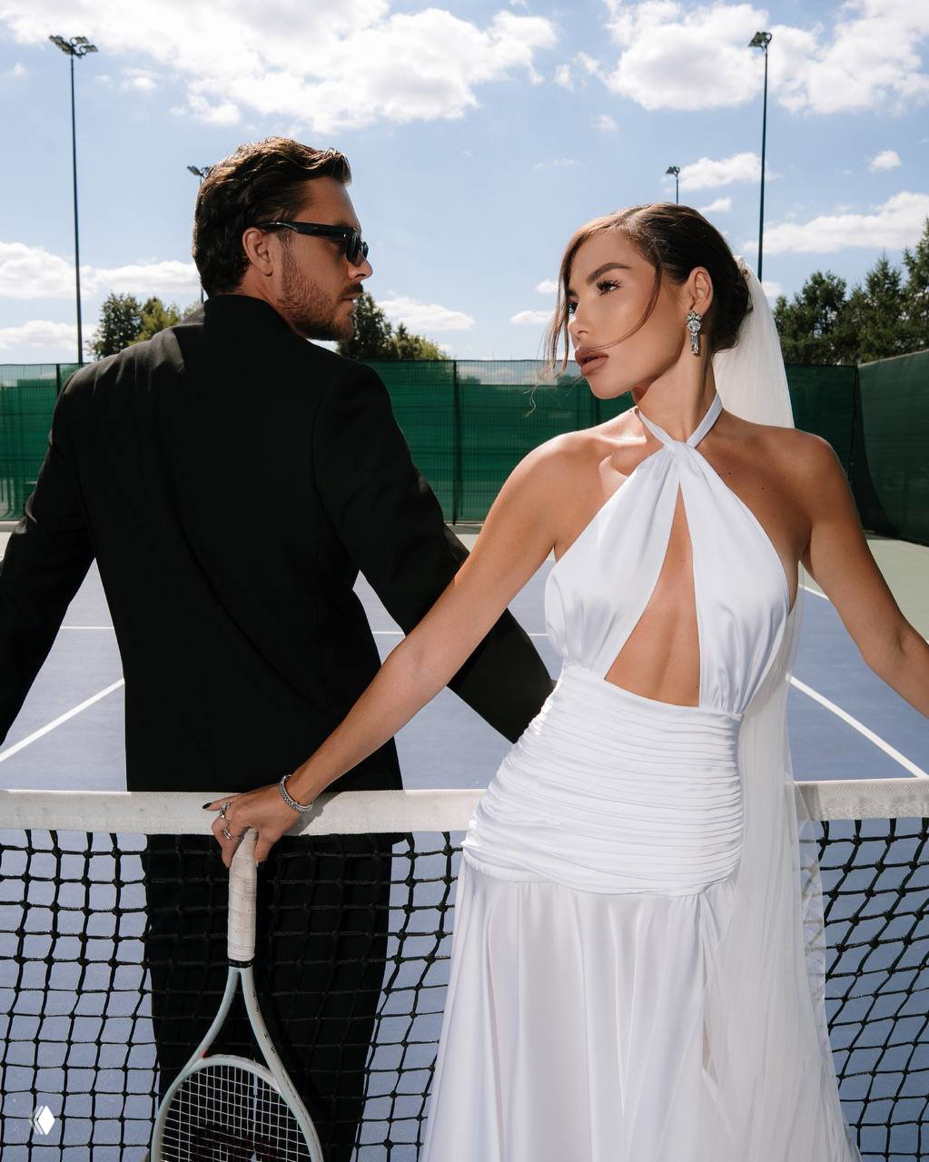 Романтичный кадр в профиль: Карина в белом платье с фатой облокачивается о сетку, рядом — мужчина в строгом костюме, зелёный фон ограждений теннисного корта.