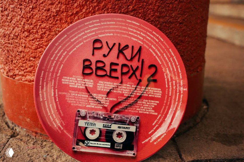 Крупный план красного диска с надписью «Руки Вверх! 2» и прозрачной аудиокассетой внизу — постерный реквизит с именами съёмочной группы