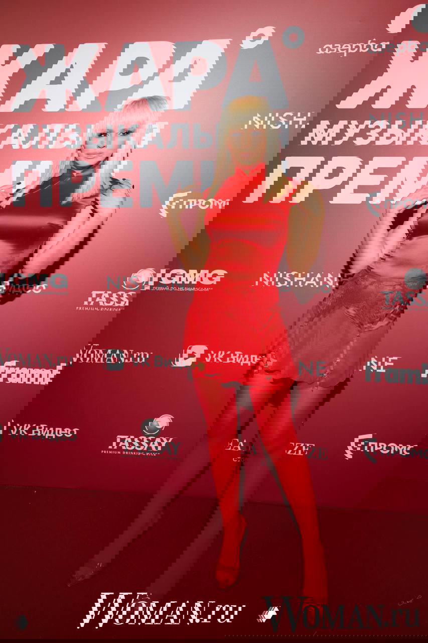 Женщина в ярком красном мини‑платье и плотных колготках позирует на красной дорожке ЖАРА; на фоне видны логотипы спонсоров и Woman.ru.