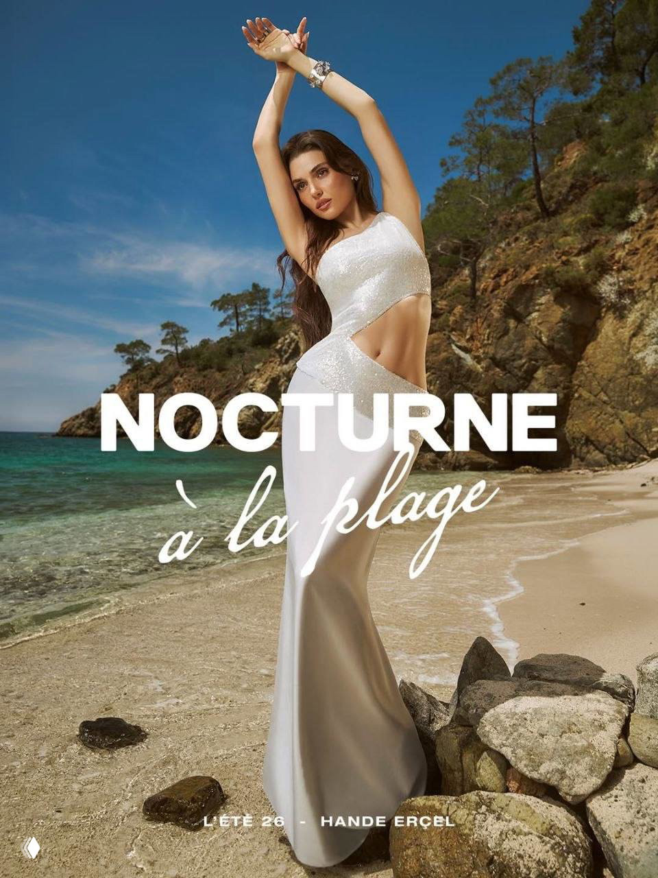 Хандэ Эрчел в длинном белом платье на пляже для кампейна Nocturne: модель позирует на берегу на фоне скал и моря, рекламный кадр.