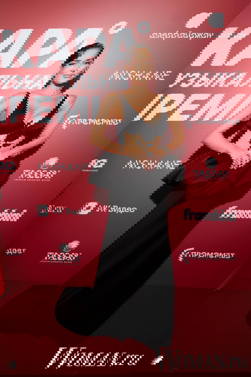 Женщина в чёрном вечернем платье с выразительным декольте и декоративным поясом позирует у фотозоны на музыкальной премии ЖАРА в Москве.