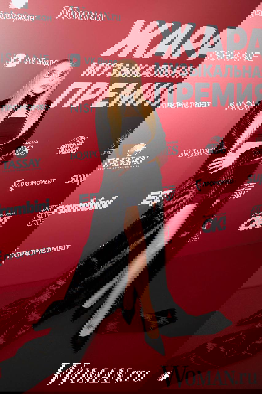 Молодая женщина в длинном чёрном бархатном плаще и коротком платье демонстрирует образ на красной дорожке ЖАРА на фоне бренд‑баннера.