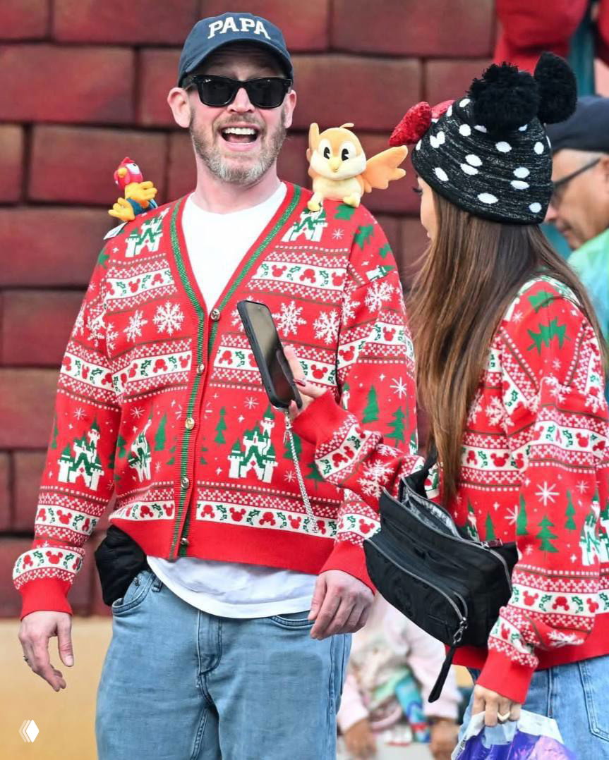 Маколей Калкин и Бренда Сонг в одинаковых праздничных кардиганах смеются и разговаривают в парке развлечений рядом с коляской.