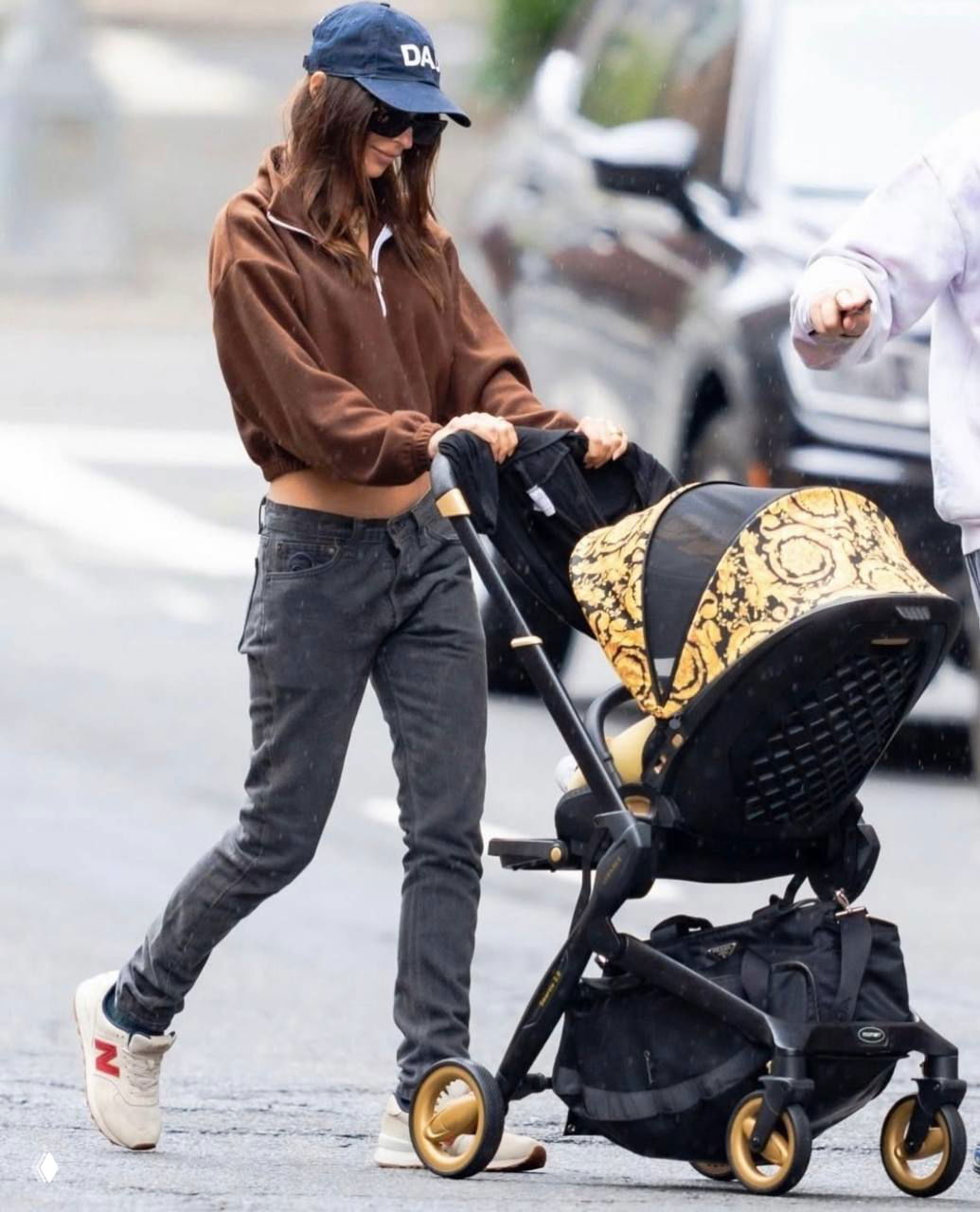 Женщина в кепке и солнцезащитных очках толкает детскую коляску с узором по городской улице — сцена о родителях и выборе имён.