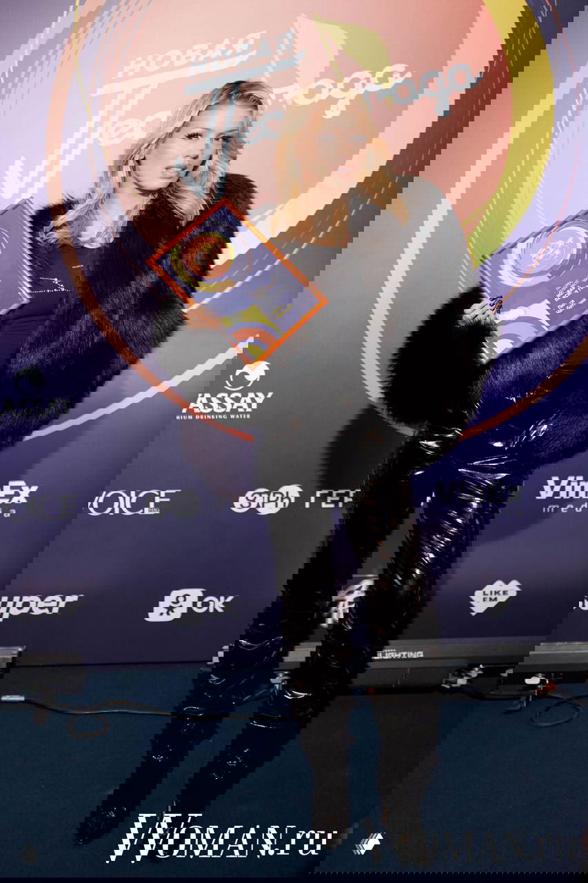 Исполнительница в чёрной шубе и лаковых брюках держит грамоту на фоне логотипа фестиваля «Новая Песня года», мероприятие 2025.