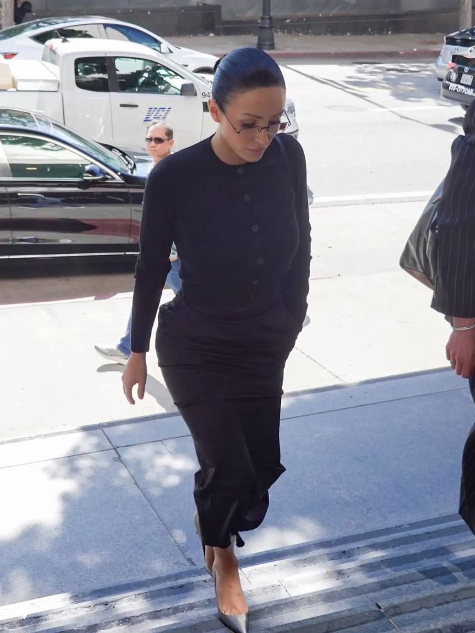 Папарацци-снимок: женщина в тёмном наряде с собранными в пучок волосами поднимается по ступеням к входу в здание.