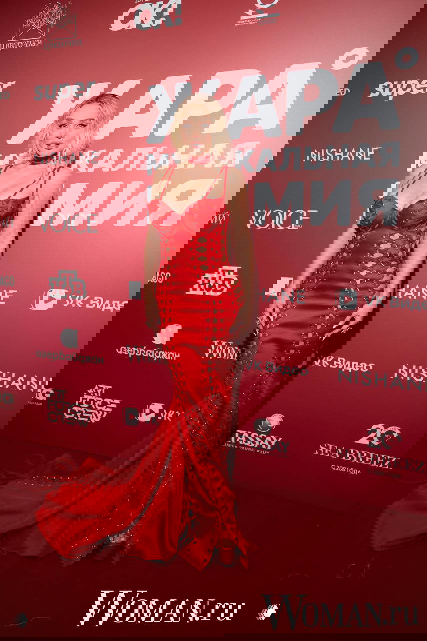 Женщина в обтягивающем красном платье со шнуровкой и разрезами позирует на красной дорожке ЖАРА перед фотофоновой панелью с логотипами.