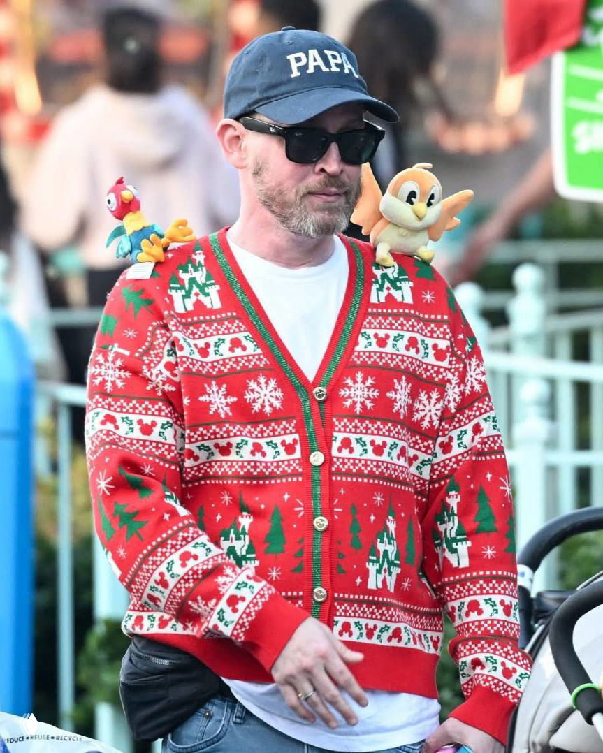 Крупный план Маколея Калкина в кепке «PAPA», тёмных очках и в красном свитере с новогодним узором и игрушками на плечах.