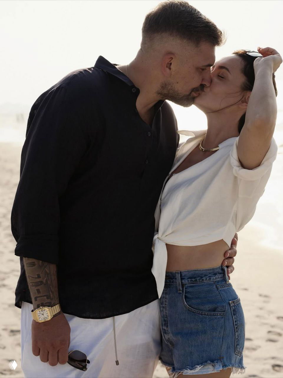 Крупный план поцелуя пары на пляже: мужчина в тёмной рубашке прижимает женщину в лёгкой белой блузе, эмоциональный момент.