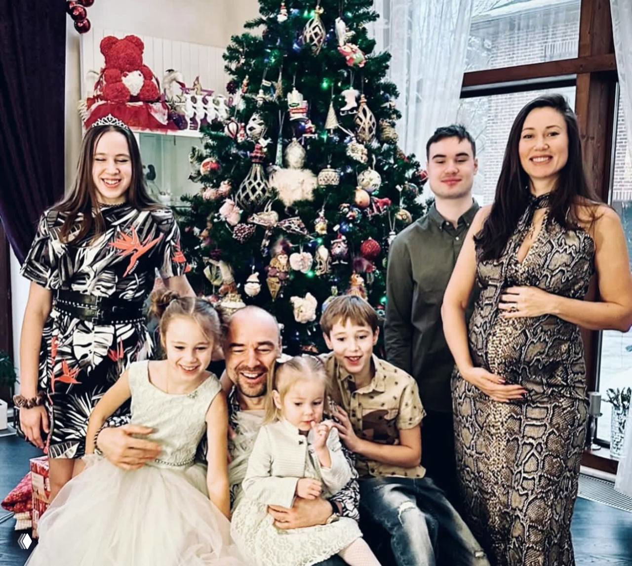 Семейная фотография у украшенной ёлки: несколько взрослых и детей позируют вместе в праздничной обстановке, видна беременная женщина.