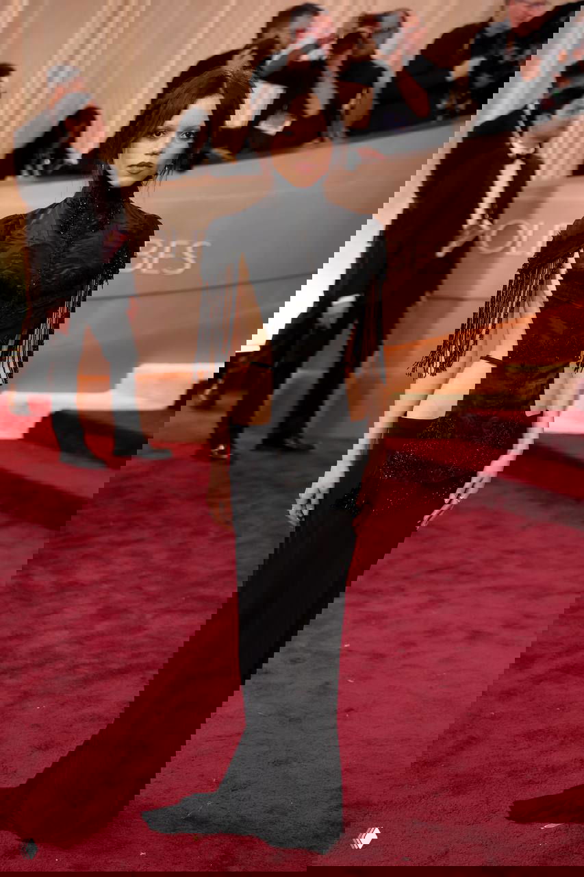 Женщина в чёрном платье с декоративными вырезами и бахромой стоит на красной дорожке, рядом папарацци и гости церемонии.