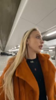 Кадр вертикального видео: блондинка в оранжевой шубе идёт по подземной парковке, смотрит в сторону, снято на ходу и динамично.