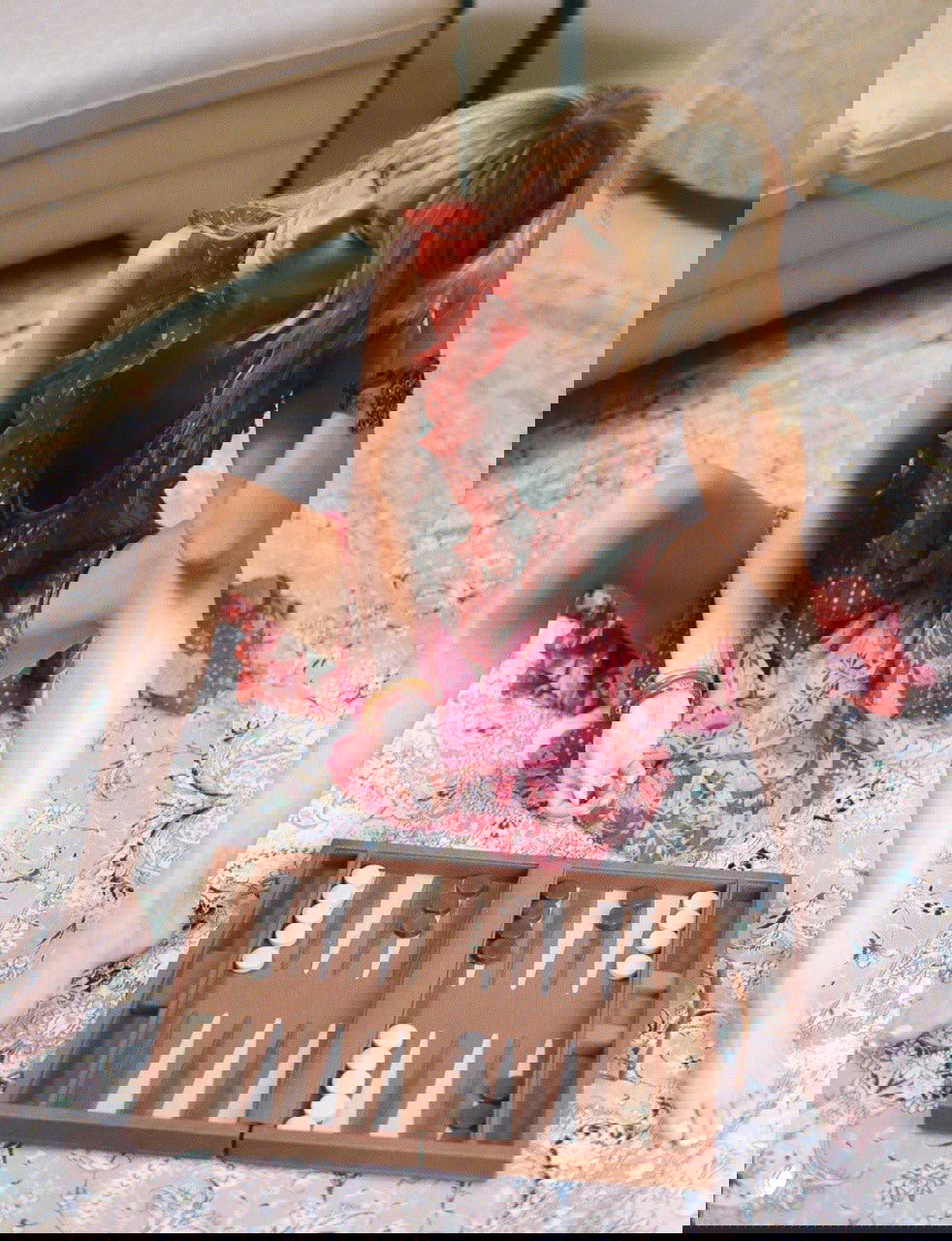 Белла Хадид в романтичном розовом платье сидит на ковре, перед ней раскладная доска для нард — непринуждённая домашняя сцена.