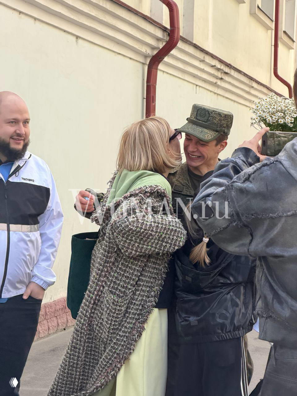 Глеб Калюжный обнимает и целует родственница при встрече: актёр в камуфляже улыбается, рядом поздравляющие и крупный букет.