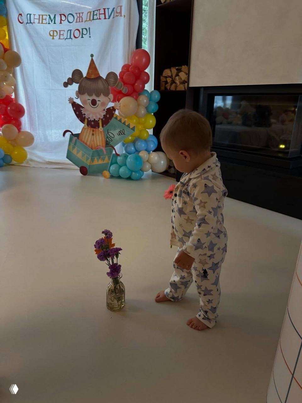 Малыш в пижаме внимательно рассматривает праздничную сцену с баннером и воздушными шарами; на полу рядом стоит стеклянная ваза с небольшим букетом цветов.