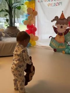 Вертикальный видеокадр: ребёнок играет с мягкой игрушкой перед декорацией «С Днем Рождения, Фёдор!», домашняя гостиная с украшениями и шариками на заднем плане.