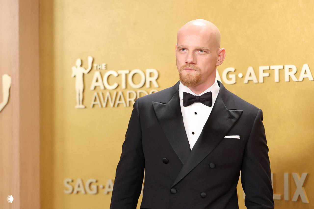 Крупный план актёра в смокинге на фоне золотой фотозоны премии SAG‑AFTRA, кадр с красной дорожки церемонии