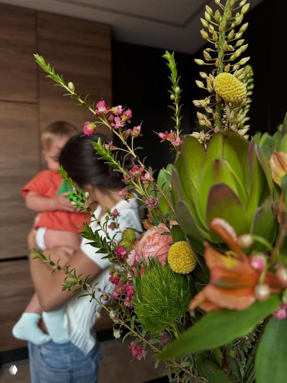 Крупный план композиции из розовых, жёлтых и зелёных цветов на переднем плане; на заднем плане размытая фигура женщины, держащей ребёнка на руках — домашний праздник.