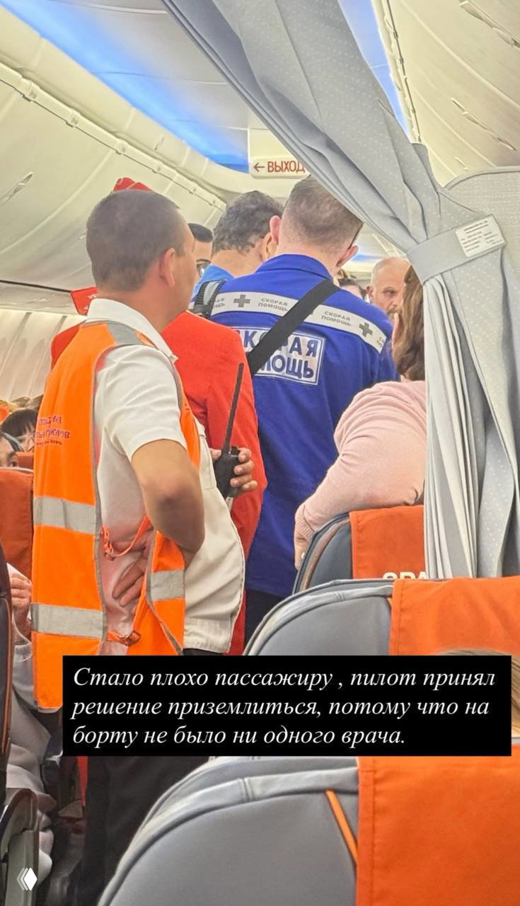 Салон самолёта: пассажиры в проходе, экипаж и медики оказывают помощь, видны оранжевые кресла и элементы интерьера самолёта.