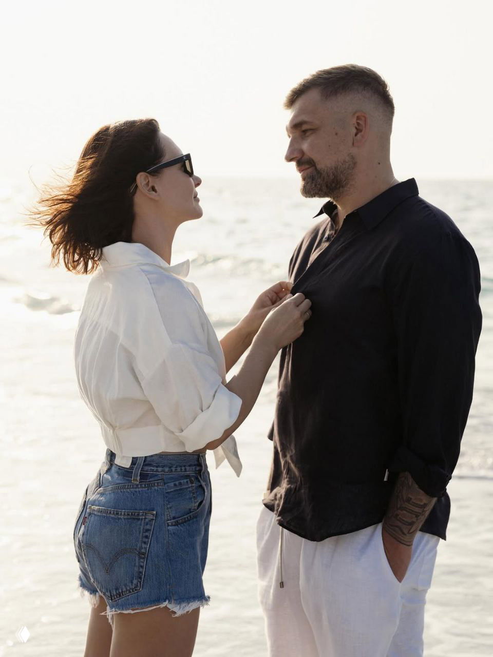 Пара стоит лицом друг к другу у воды: женщина поправляет мужчине рубашку, лёгкий ветер раздувает волосы и одежду.