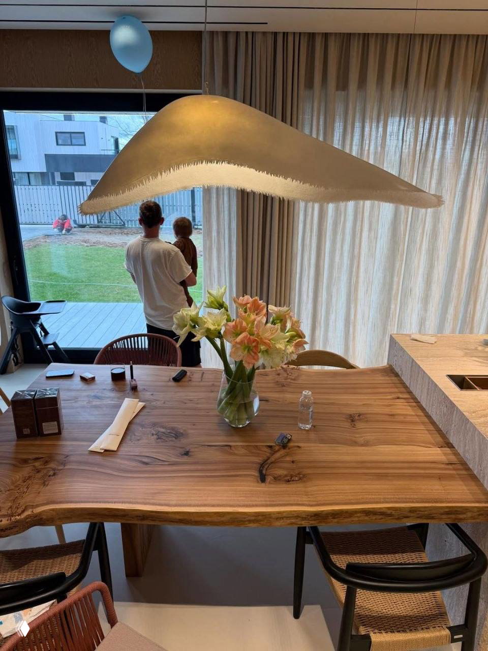 Папа держит сына у большого панорамного окна кухни: на столе ваза с цветами, виднеется терраса и зелёная лужайка во дворе дома — семейная сцена после праздника.