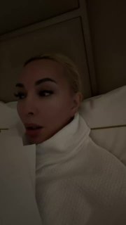 Фрагмент видео: женщина в светлой одежде лежит или сидит на кровати, лицо в полупрофиле, спокойный интерьер и приглушённый свет.