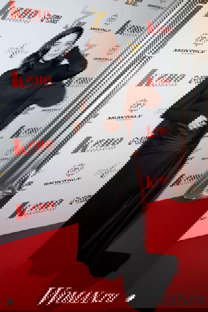 Женщина в длинном темном платье позирует на фоне пресс-волла «Кино Репортер», акцент на силуэте и глянцевом материале наряда.