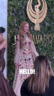 Женщина в длинном светлом платье с цветочной вышивкой на дорожке у зелёного фона с эмблемой премии Joy Awards и надписью HELLO