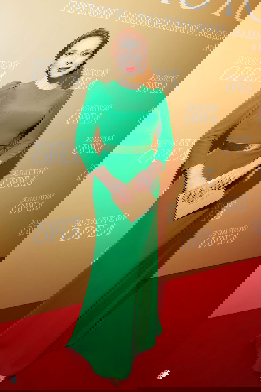 Екатерина Гусева в элегантном зеленом платье с поясом и клатчем позирует перед фотозоной премии «Золотой орёл» на красной дорожке.