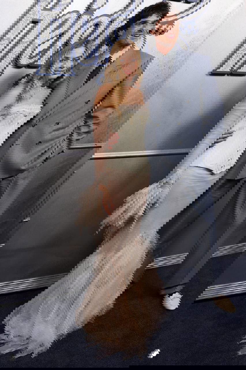 На фото пара на премьере: женщина в светлом платье и мужчина в двубортном костюме позируют у декоративной стенки с логотипом фильма.