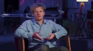 Кадр из студийного интервью: мужчина в светлой рубашке сидит в кресле и жестами объясняет на тёмном фоне