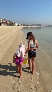 Пляжный кадр: женщина с двумя детьми идёт по песчаному берегу, следы на песке и спокойная прибрежная вода на фоне застройки.