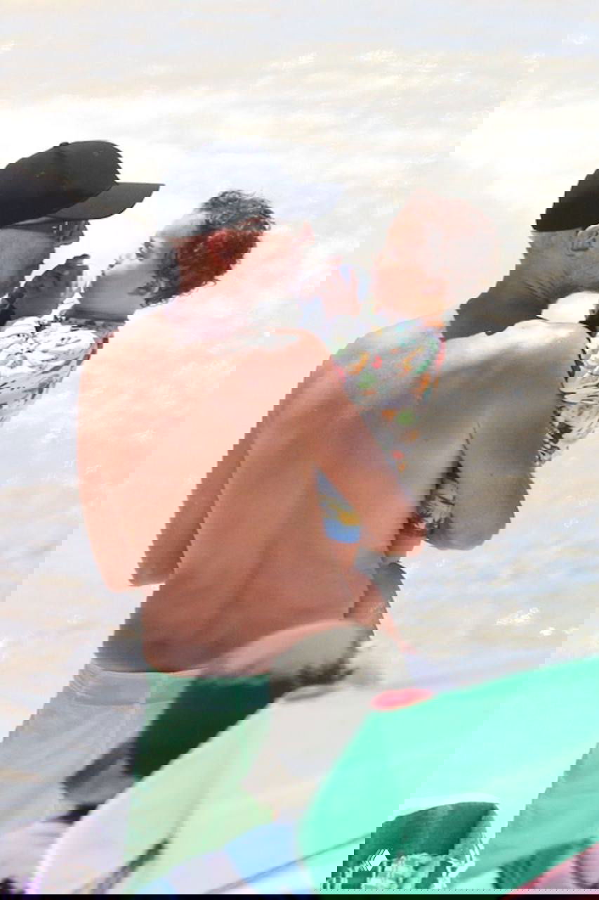 Мужчина держит на руках маленького ребёнка у воды; кадр снят со спины, видно солнце, прибой и пляжную атмосферу.