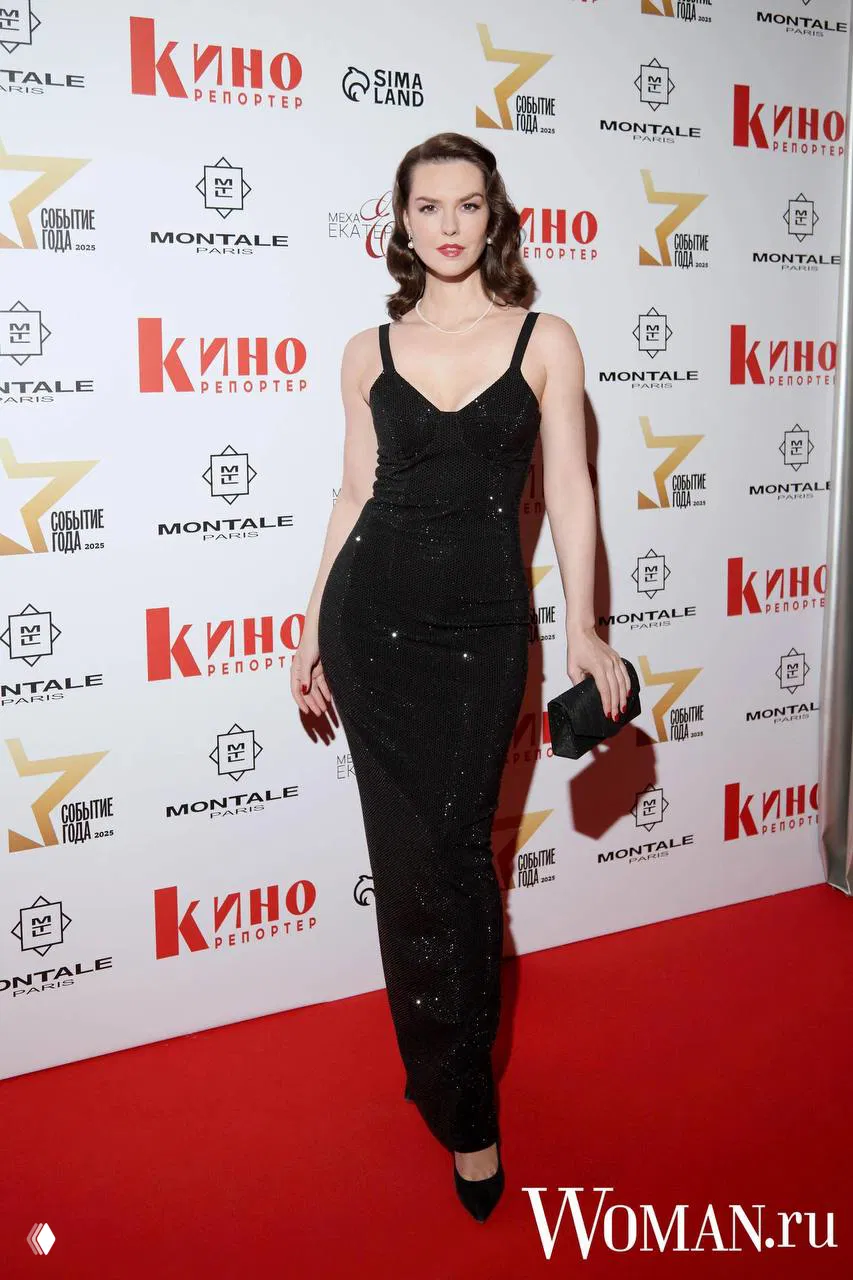 Женщина в чёрном длинном платье с открытыми плечами и жемчужным украшением, полный рост на красной дорожке у баннера мероприятия.