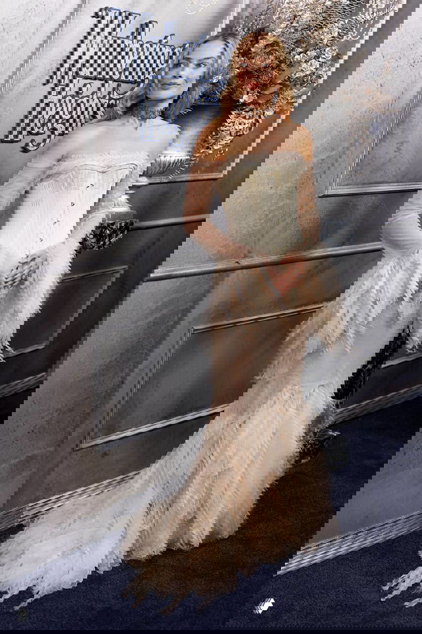 Полноформатный портрет женщины в воздушном жемчужно-кремовом платье и корсете на фоне изысканных декораций и логотипа премьеры.