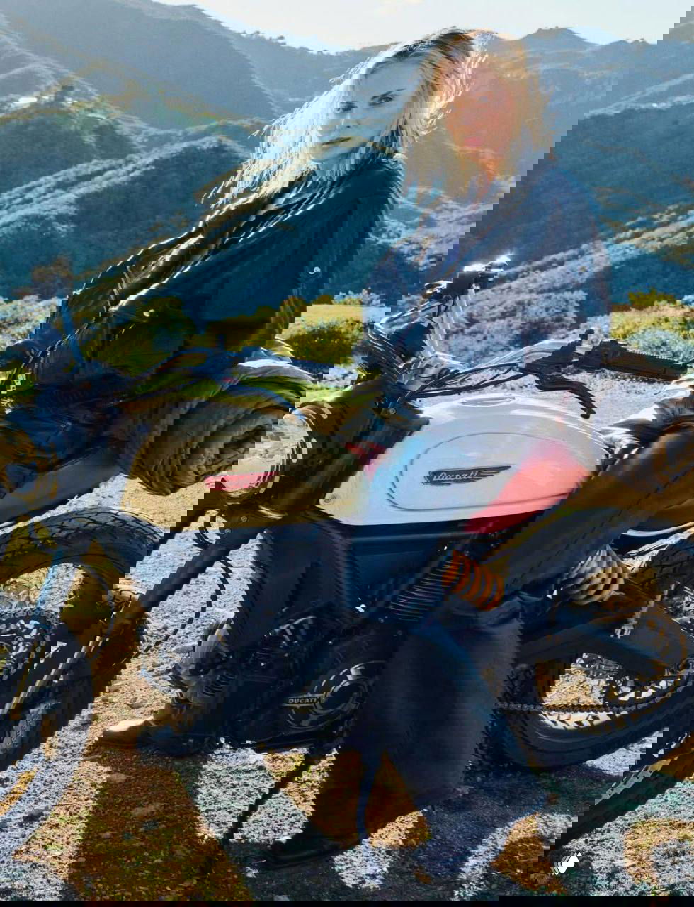 Женщина в тёмной кожаной куртке сидит на мотоцикле на фоне холмов; полный рост, видна часть мотоцикла и дорога.