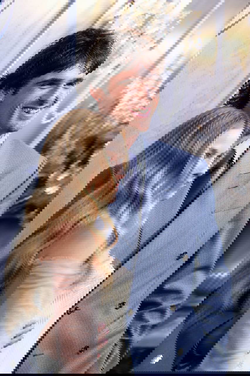 Крупный план улыбающегося мужчины в голубом костюме рядом с женщиной в корсете; на заднем плане серебристая фотозона премьеры фильма.