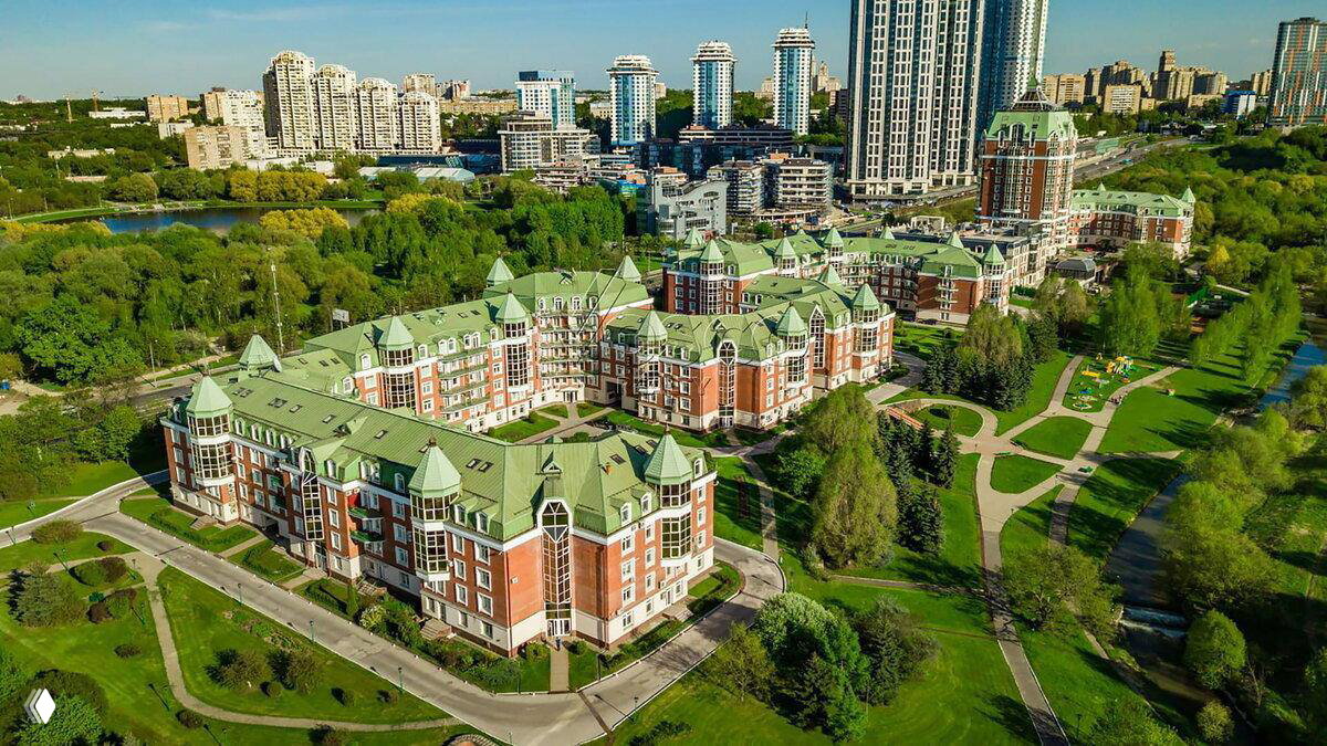 Аэрофотоснимок жилого комплекса и прилегающего парка: кирпичные корпуса с зелёными крышами, дорожки и окружающая городская застройка с зелёными зонами.