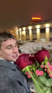 Мужчина в зимнем пальто держит большой букет красных и розовых цветов на улице на фоне снежных сугробов и зданий.