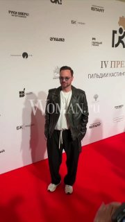 Мужчина в тёмной куртке и светлой рубашке стоит у бренд‑стены на красной дорожке — момент с фотосессии на церемонии премии кастинг‑директоров.